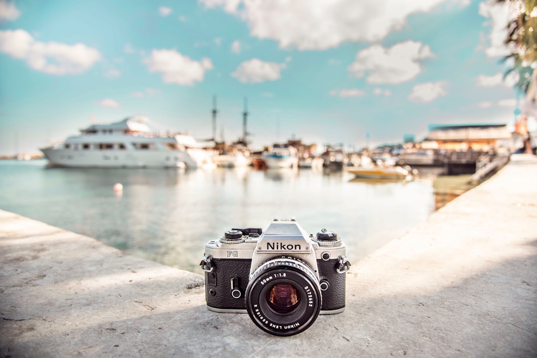 A Nikon film camera sits on a concrete surface near a marina, with luxury boats and blue sky with clouds in the blurred background, evoking the allure of private yacht tours in the Caribbean.