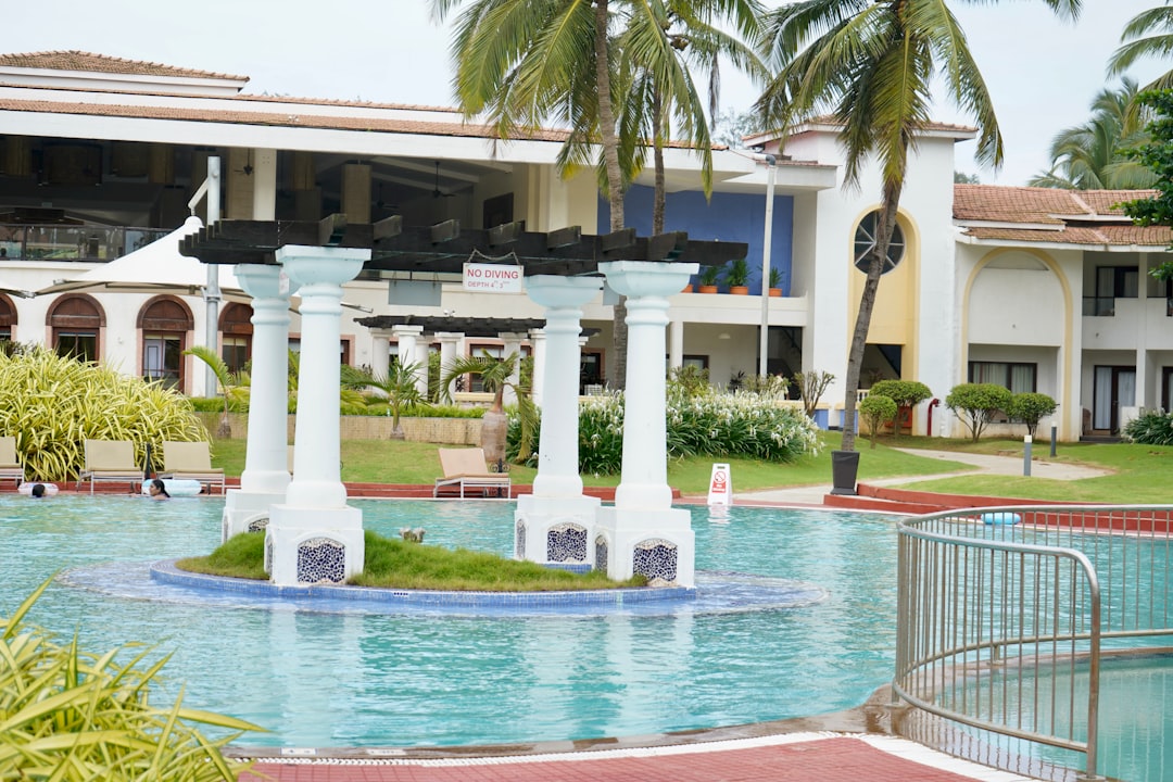 Outdoor swimming pool with white columns on a small island, surrounded by lounge chairs, palm trees, and a two-story resort building—perfect for relaxing after a luxury helicopter tour.
