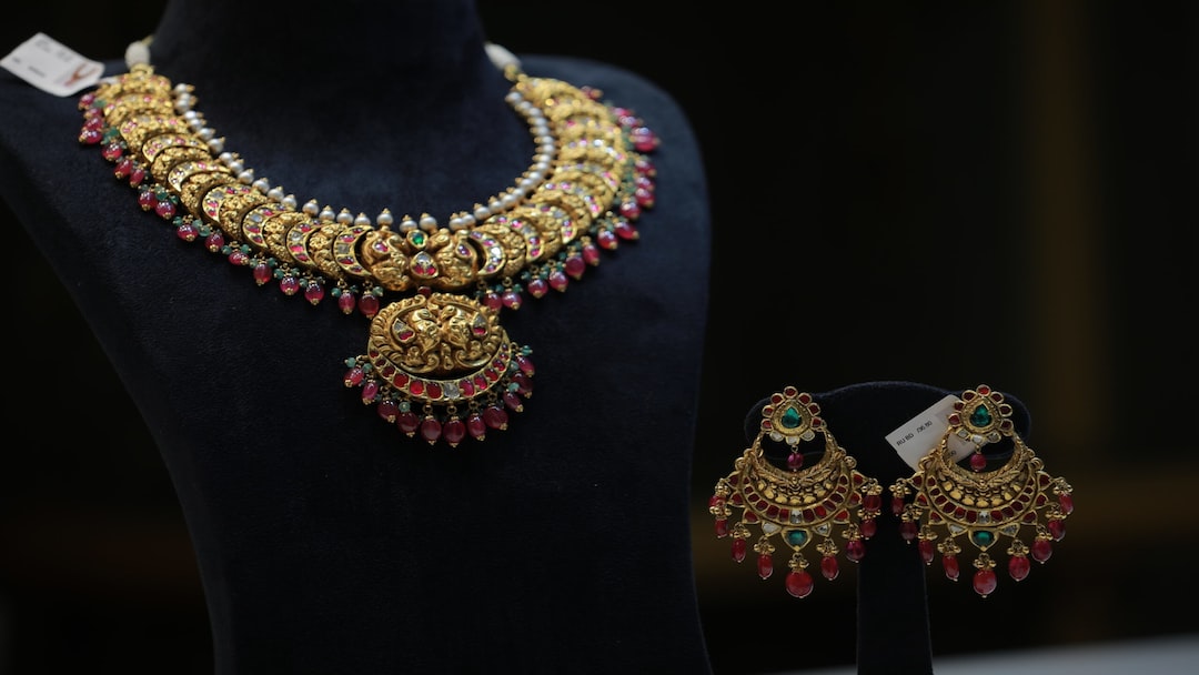 A gold necklace with red, green, and white gemstones displayed on a black stand, accompanied by matching earrings on a separate smaller stand in the heart of London's Diamond District.