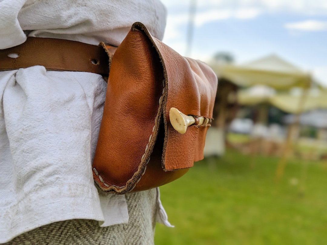 Close-up of a brown leather pouch with a bone button, showcasing skilled craftsmanship, attached to a belt worn over a white shirt and grey pants, outdoors.