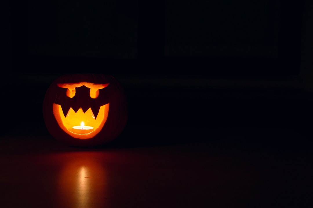 A carved pumpkin with a menacing face is lit from within by a candle, casting a glow in a dark room reminiscent of fall beauty.