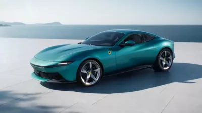 A luxury teal Ferrari coupe is parked on a paved surface, with an ocean and distant islands in the background under a clear sky.