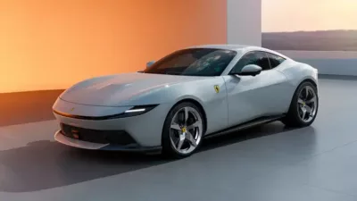 A silver Ferrari luxury sports car is parked indoors on a smooth floor, with an orange-lit background and large windows revealing an outdoor scene.