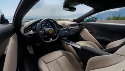 Interior view of a modern Ferrari car showcasing a black luxury steering wheel with the Ferrari logo, digital dashboard, and beige leather seats, all set against a stunning coastal landscape visible through the windows.