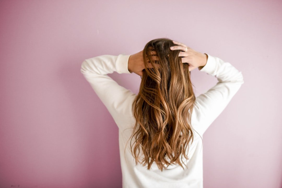 Person with long, wavy brown hair wearing a white long-sleeve shirt, facing a light purple wall with hands on the back of their head, embracing flawed beauty and the charm of Schönheitsfehler.