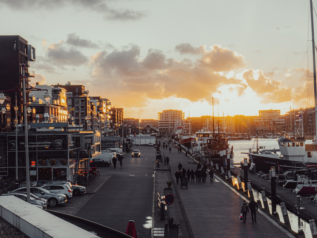 People stroll along a harbor promenade lined with modern buildings and luxury yachts at sunset, with cars parked on the left and the sky partly cloudy.