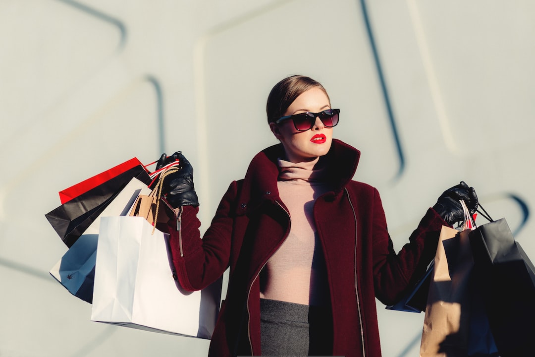 A woman wearing sunglasses and a burgundy coat holds multiple shopping bags in both hands against a light gray background, channeling the bold spirit of experimental fashion.