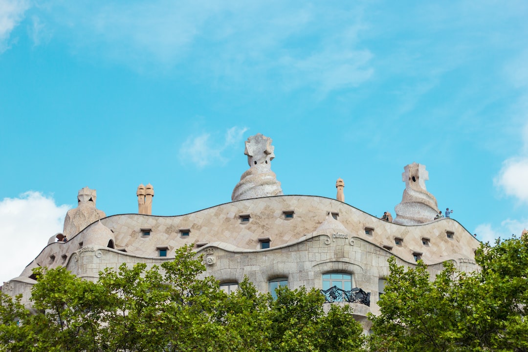 Rooftop of an ornate stone building with sculptural chimneys and wavy lines, partially obscured by green trees, under a bright blue sky—a signature Gaudi masterpiece in Barcelona ideal for luxury tours.