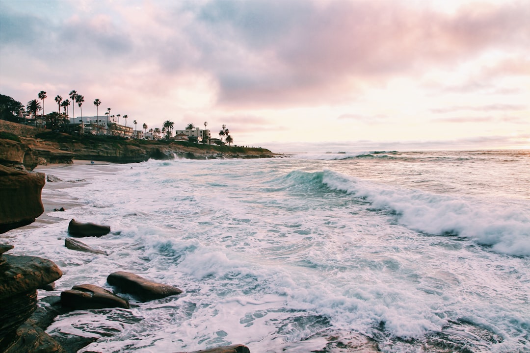 Waves crash effortlessly against rocky cliffs at the coastline, with palm trees and buildings visible in the distance under a cloudy sky at sunset.