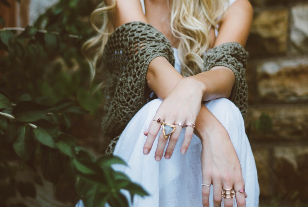 A person with long blonde hair wearing iconic jewelry, a white dress, and a green knitted shawl sits on outdoor stone steps with green leaves in the foreground.