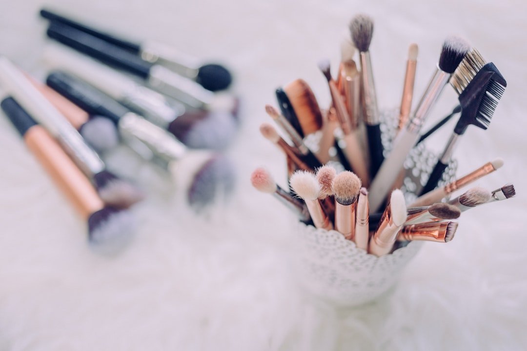 A collection of makeup brushes and applicators in a holder with more brushes lying on a white, fluffy surface in the background, inspired by Global Beauty Secrets and Beauty Traditions from around the world.