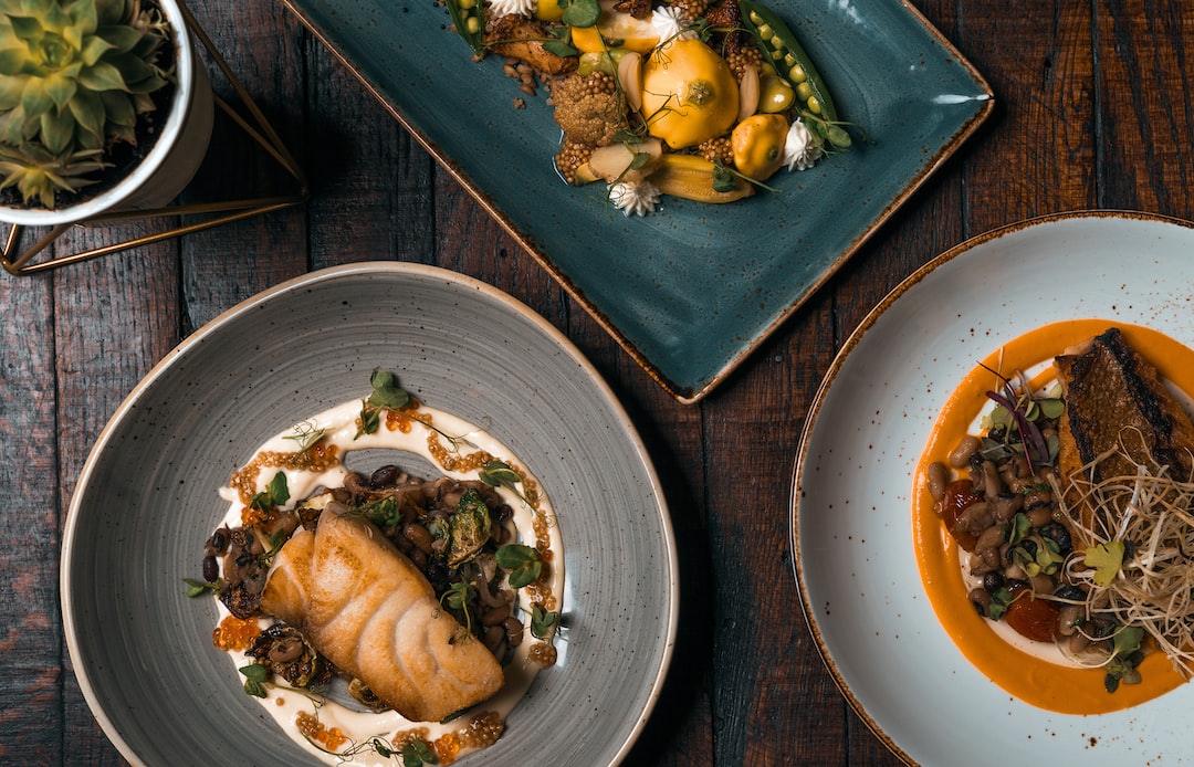 Three plated gourmet dishes—fish, vegetables, and a meat entrée—are artfully arranged on a wooden table, echoing the refined style of Gourmet Berlin and capturing the essence of luxury dining with a small potted plant in the top left corner.