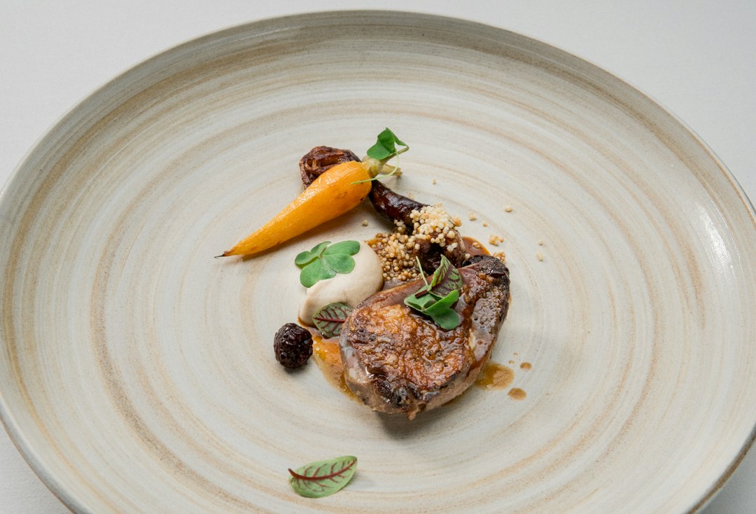 A plated dish at one of the best restaurants San Francisco offers, featuring seared meat, a small carrot, a dollop of puree, garnish, and sauce—artfully arranged on a large beige ceramic plate for an unforgettable fine dining experience.