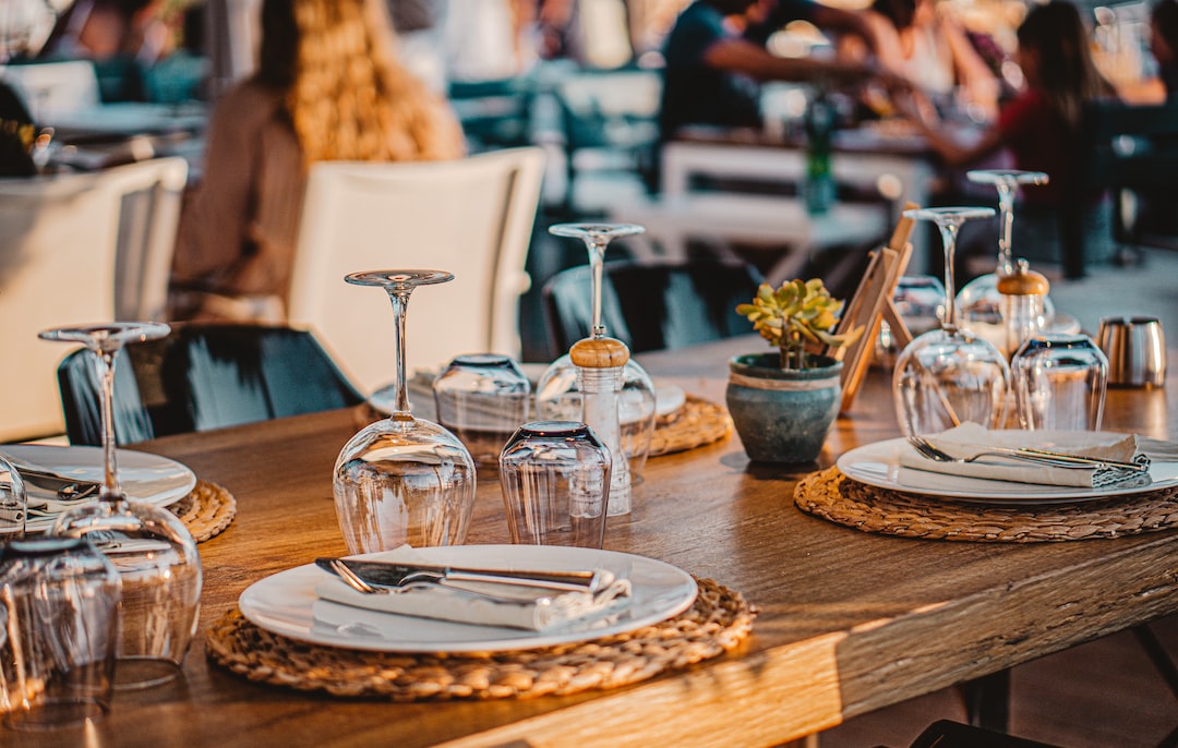 A wooden table is set with plates, cutlery, napkins, upside-down wine glasses, and a small potted plant in an exclusive dining outdoor LA setting with people mingling in the background.