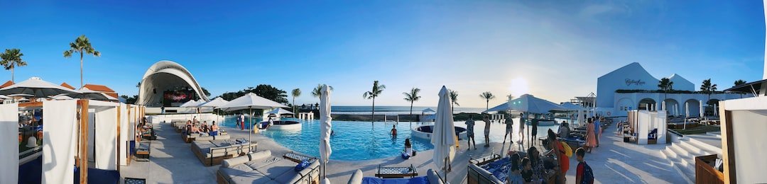 Panoramic view of a resort pool area with lounge chairs, cabanas, umbrellas, and palm trees by the ocean under a clear blue sky at sunset, reflecting the relaxed elegance inspired by Gourmet Melbourne and Australia’s Culinary Innovations.