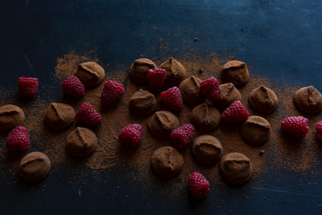Chocolate truffles dusted with cocoa powder are scattered on a dark surface alongside several fresh raspberries, evoking the indulgence of gourmet Zurich chocolates.