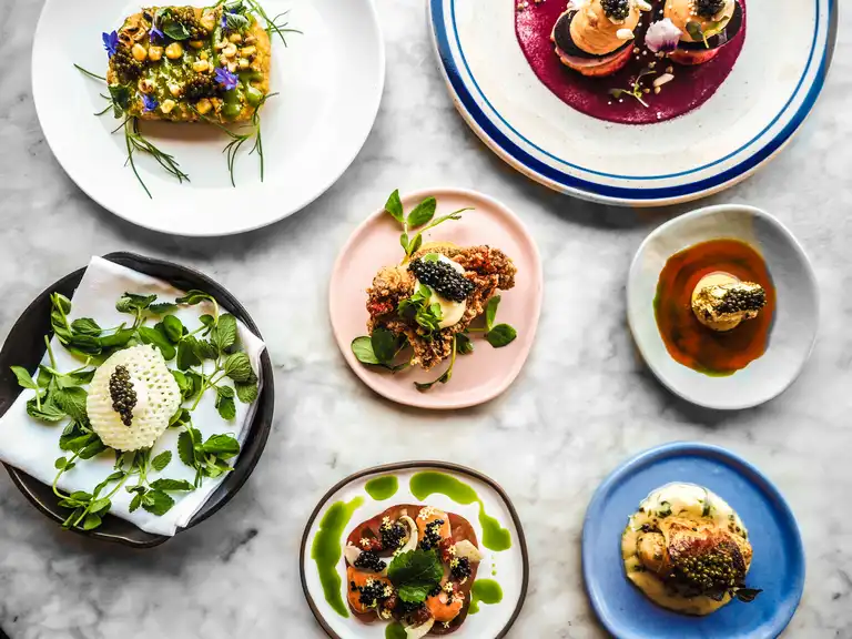 Seven artfully plated luxury gourmet dishes featuring ingredients like caviar, herbs, and colorful sauces are arranged on a marble surface.