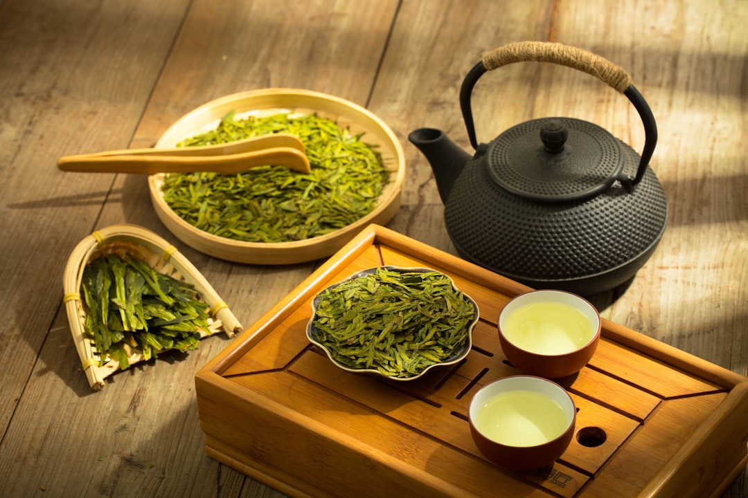 A teapot, two cups of green tea, and bowls of loose leaves are arranged on a wooden surface in natural light, showcasing green tea as a natural elixir with beauty benefits.