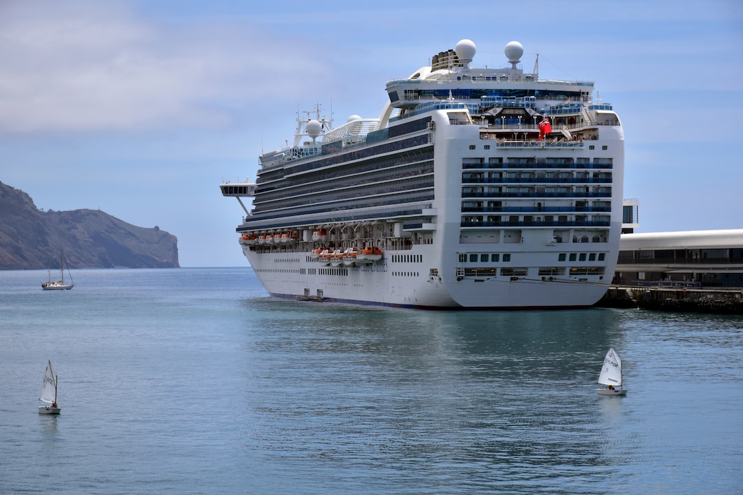 A large white luxury cruise ship docked at Victoria Harbour, with small sailboats in the water nearby and a rocky coastline in the background.