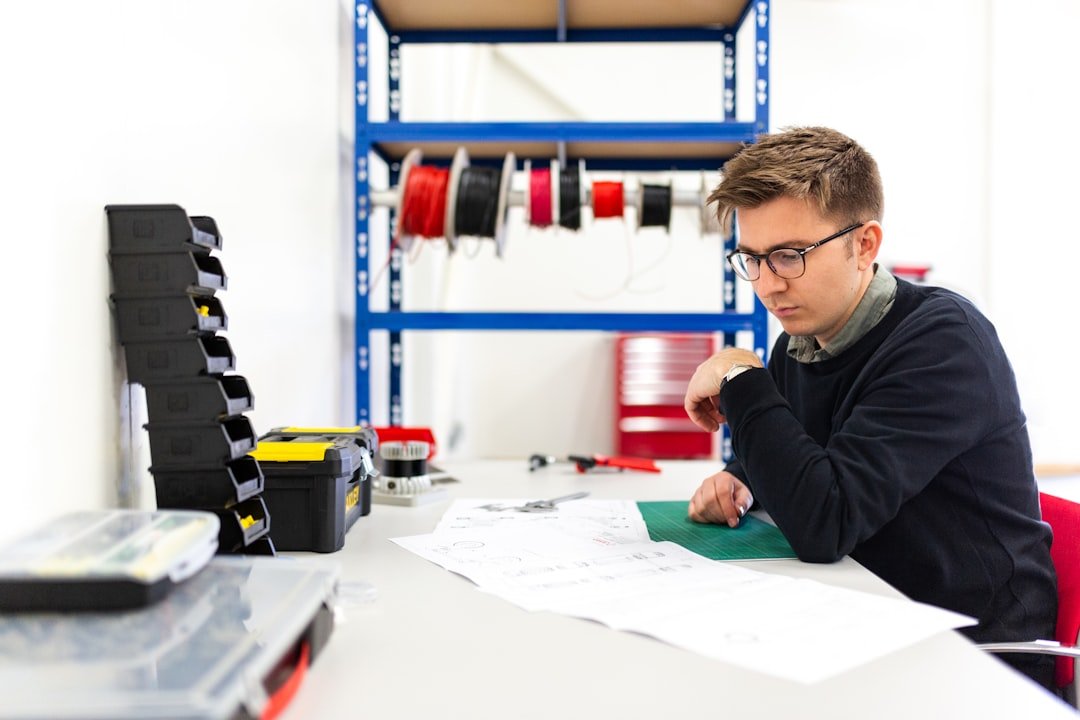 A man sits at a desk examining technical drawings, with organized tools and spools of wire on shelves in the background, his healthy nails reflecting careful attention to detail.