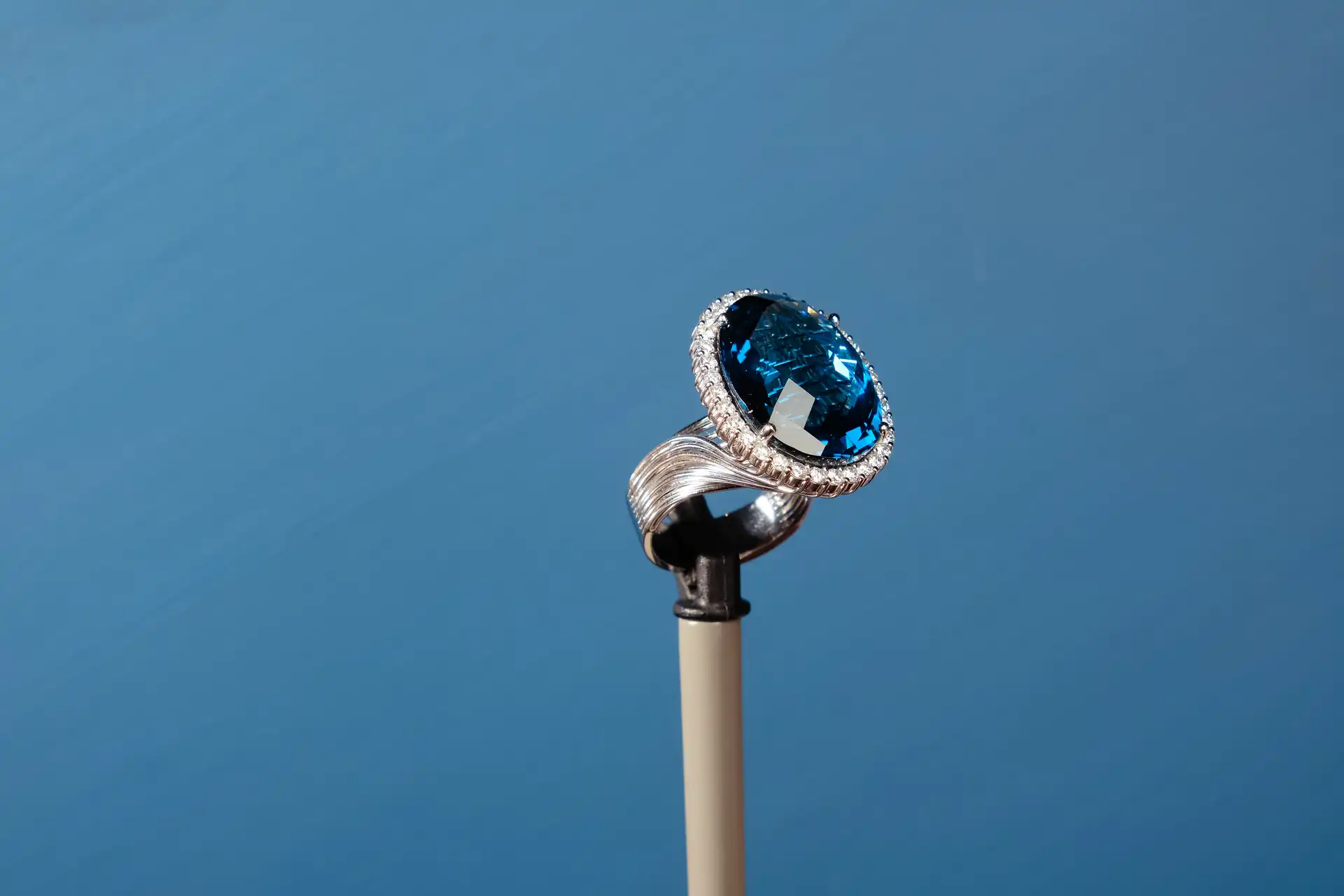 A luxury silver ring with a large oval blue gemstone and small clear stones around it is displayed on a stand against a blue background.