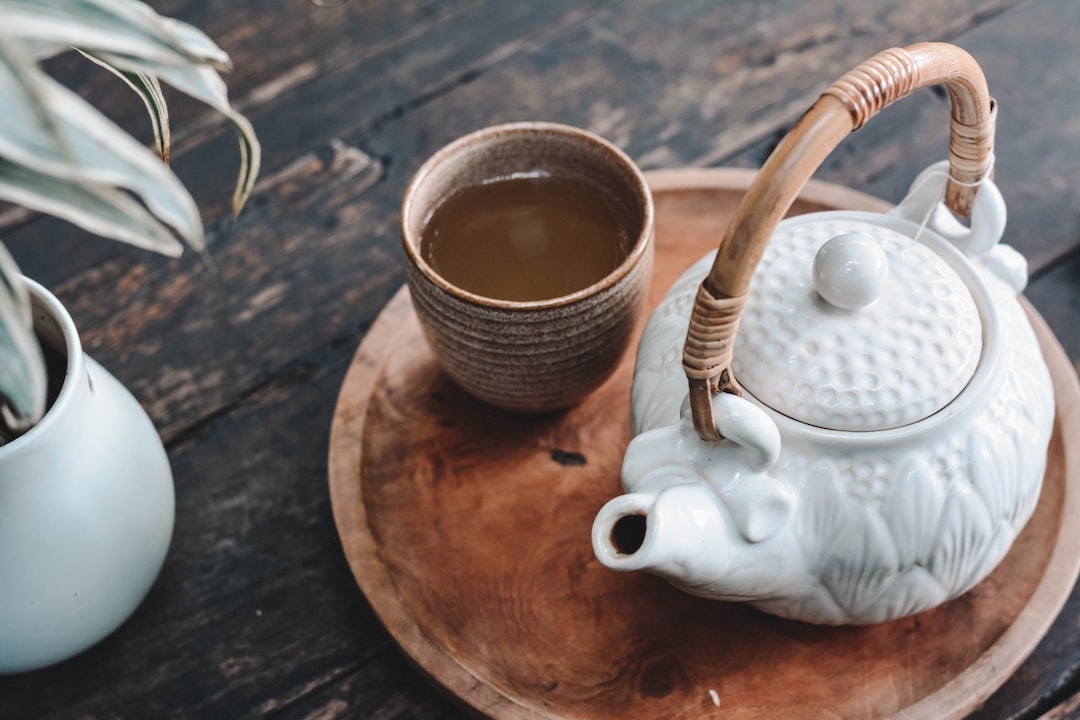 A white ceramic teapot and a cup of tea sit on a round wooden tray atop a rustic wooden table, with a potted plant nearby—perfect for enjoying a luxury afternoon tea at home.