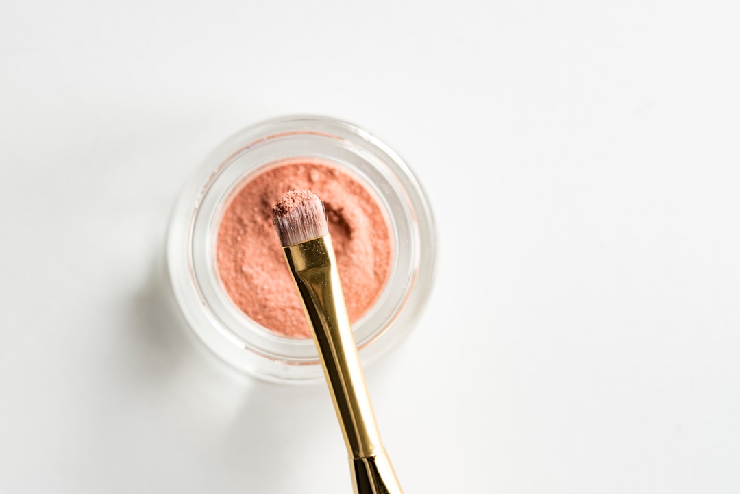 A makeup brush with a gold handle rests on the rim of a small jar filled with loose pink powder, perfect for flawless foundation application, set against a white background.