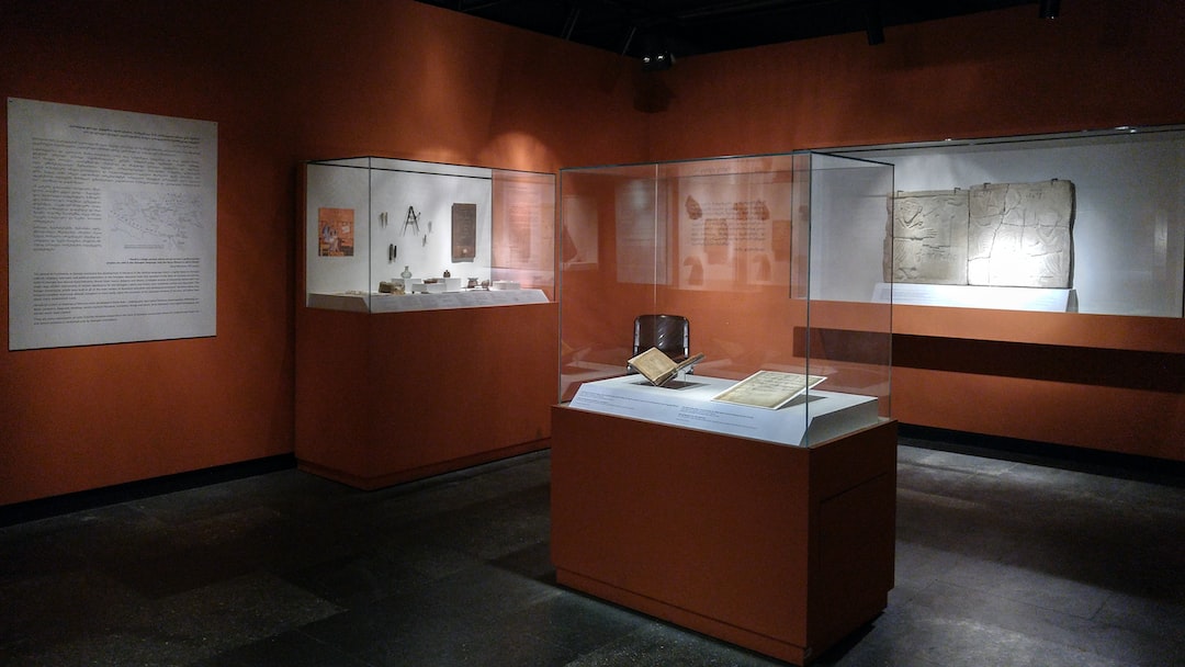 Museum room with orange walls displaying historical artifacts and iconic jewelry in glass cases, including documents, objects, and stone tablets, all showcased under focused lighting.