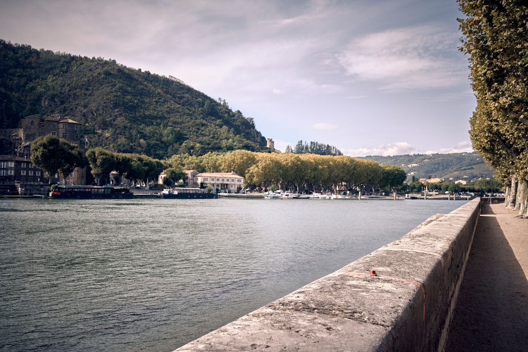 A riverside walkway with trees on the right, a stone wall, boats on the water, and wooded hills in the background under a partly cloudy sky—perfect scenery for one of our customized wine tours France or delightful cheese tours France.