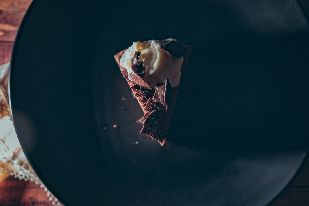 A slice of chocolate cake topped with whipped cream and chocolate shavings on a dark plate, photographed from above—the perfect indulgence for those who appreciate luxurious culinary journeys.