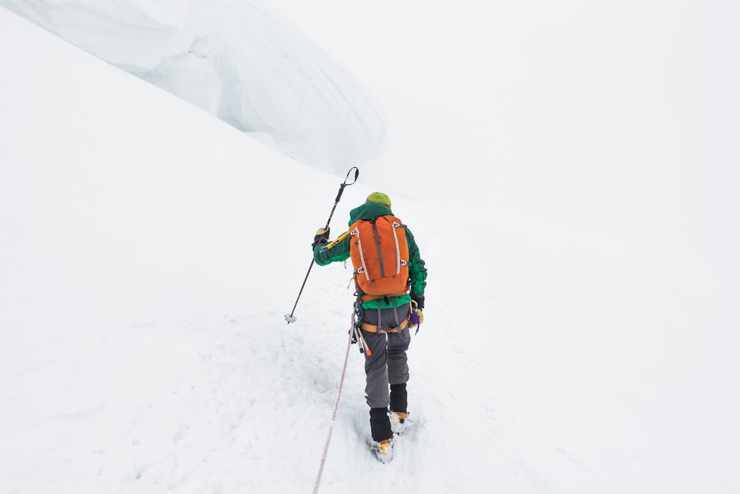 A climber wearing winter gear and an orange backpack hikes up a snowy, foggy slope using an ice axe, embracing trekking adventures on a journey to remember.
