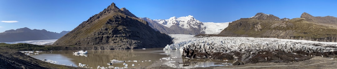 Panoramic view of a glacier flowing into a muddy lake surrounded by rocky mountains under a clear blue sky—an unforgettable sight often featured on luxury tours in Iceland.
