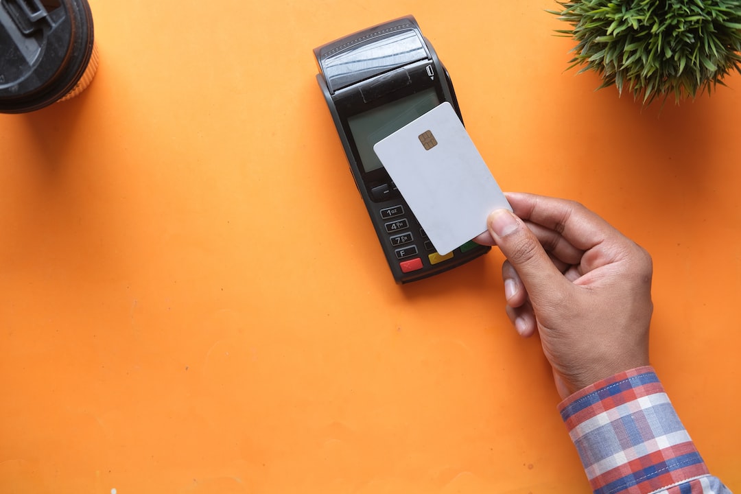A person holds a white chip card near a card payment terminal on an orange surface, ready for some retail therapy, with a plant and coffee cup nearby.