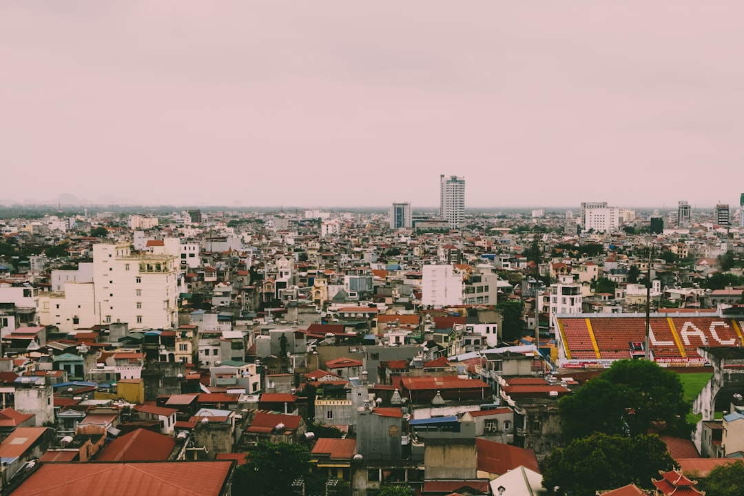 A wide view of a densely populated city with many low-rise buildings and red rooftops, a stadium to the right, and taller buildings in the distance under an overcast sky, perfect for spotting routes of Luxury Train Travel Asia.