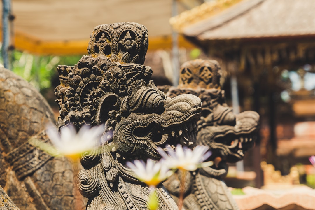Close-up of intricately carved stone statues featuring dragon-like faces, with blurred flowers and traditional structures in the background, capturing the spirit of Bali paradise near luxurious resorts Bali.