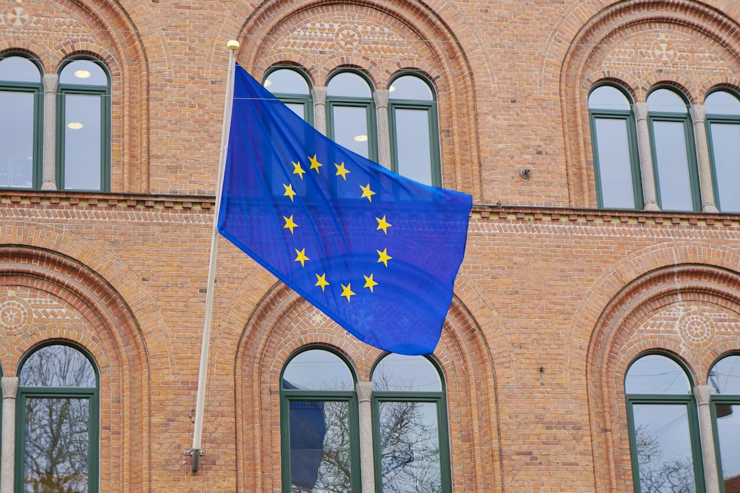 A European Union flag with yellow stars on a blue background is mounted on a pole in front of a brick building with arched windows, evoking the timeless elegance found in castle hotels across Europe.