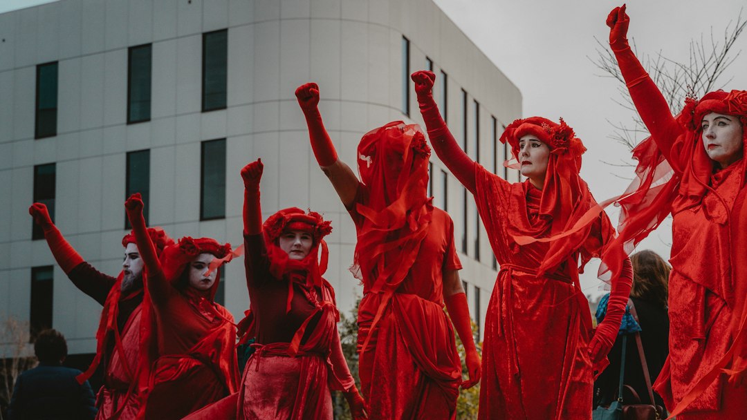 Six people dressed in red costumes and face paint stand with raised fists in front of a modern building, participating in a public demonstration advocating for sustainable luxury and eco-resorts.