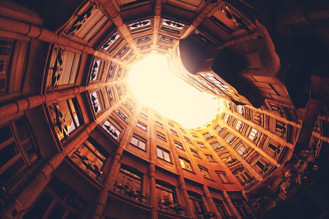 Looking up through a circular courtyard in Barcelona, surrounded by multiple stories of a building, with bright sunlight streaming through the opening above—a perfect city break moment.