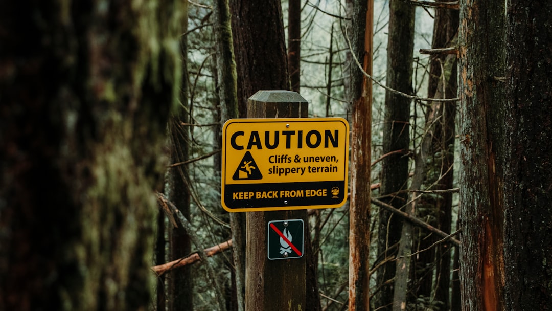 A yellow caution sign warns of cliffs and slippery terrain—advising to keep back from the edge—with a no pets sign posted below it on a wooden post among trees, reminiscent of scenic views seen on Canada train trips.