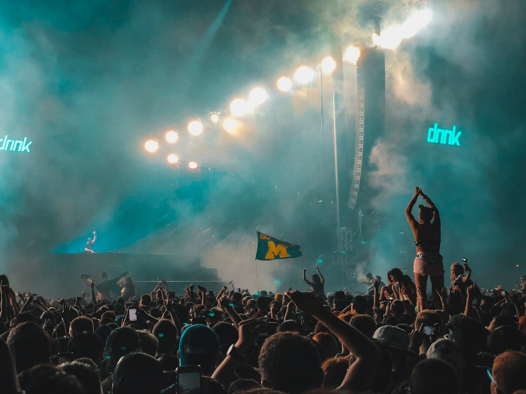 A large crowd at a nighttime concert, with stage lights illuminating the smoke-filled air; a person on someone’s shoulders waves a flag with a yellow "M" on it, capturing the energy found in luxury desert resorts.