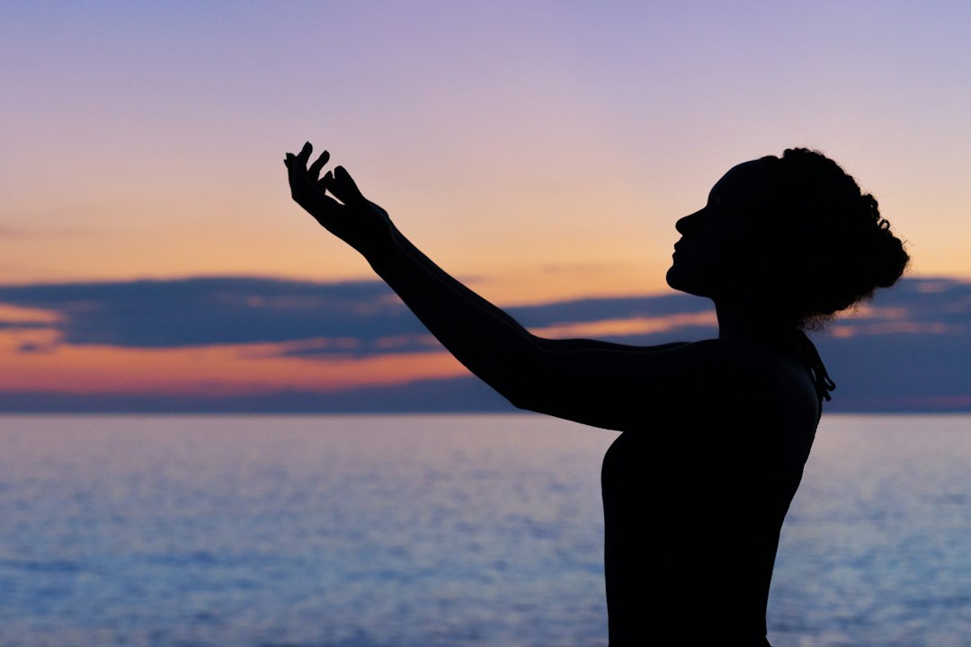 Silhouette of a person with raised arms standing by the sea at sunset, capturing the essence of luxurious relaxation and tranquility often found at wellness retreats, set against a vibrant, colorful sky.