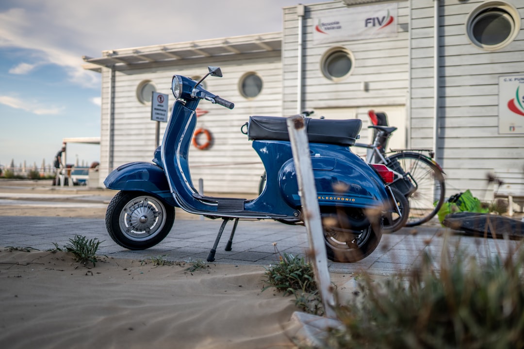 A blue vintage scooter parked on a paved path near a modern white building with round windows evokes the charm of Italy's finest cuisine, while a bicycle rests in the background.