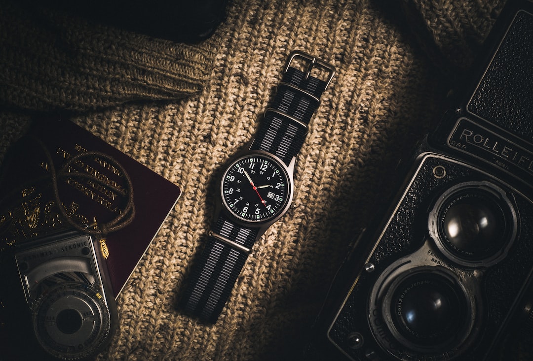 A black wristwatch with a striped nylon strap is placed on a knitted surface, surrounded by a camera, a passport, and an exposure meter—a classic scene for vintage watches collectors or those interested in investing.
