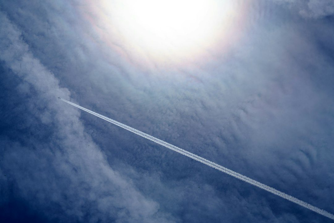 A jet plane flies through a blue sky, leaving a white contrail behind it as it heads toward luxurious getaway destinations, with the sun shining brightly above.