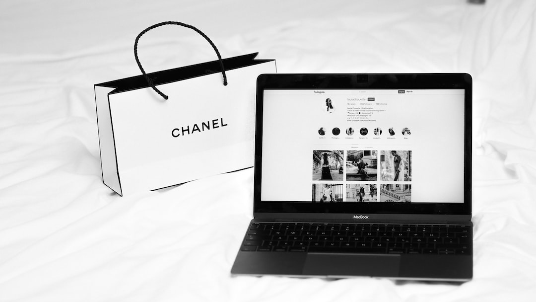 A Chanel shopping bag and an open MacBook displaying an Instagram profile are placed on a white bed, capturing the essence of a fashion brand lifestyle.