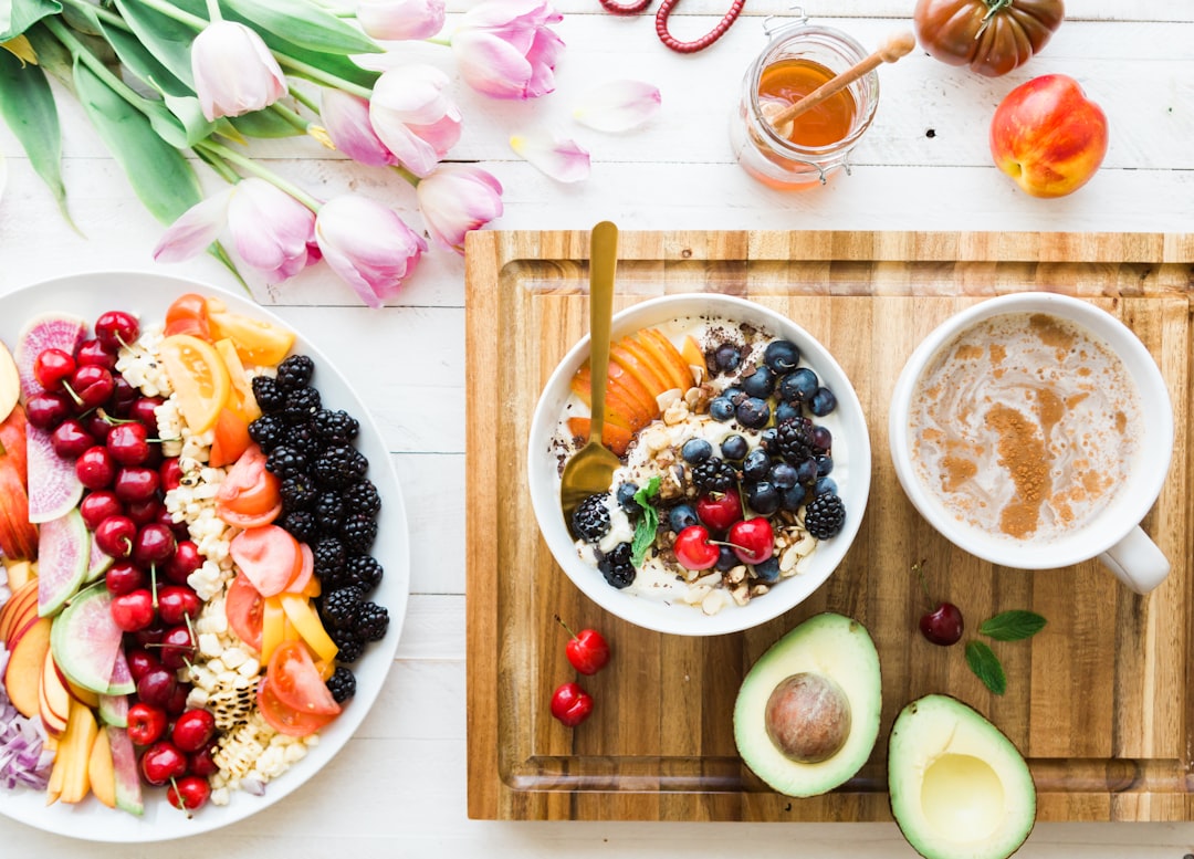 A wooden tray with a bowl of fruit and granola, a cup of coffee, and an avocado; next to a plate of assorted fresh fruits and granola, flowers, and a jar of honey—perfect for relaxation at luxurious retreats.
