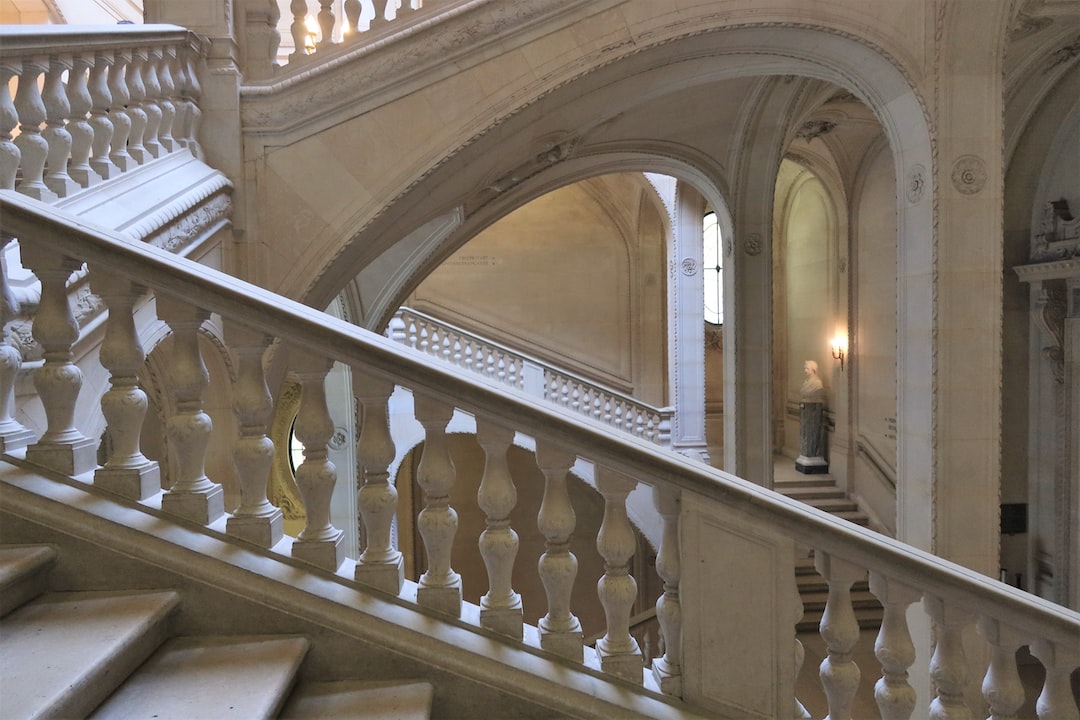 Grand stone staircase with ornate balustrades, arches, and a statue in a classical interior with soft lighting—an exquisite showcase of Milanese Tradition and refined furniture design.