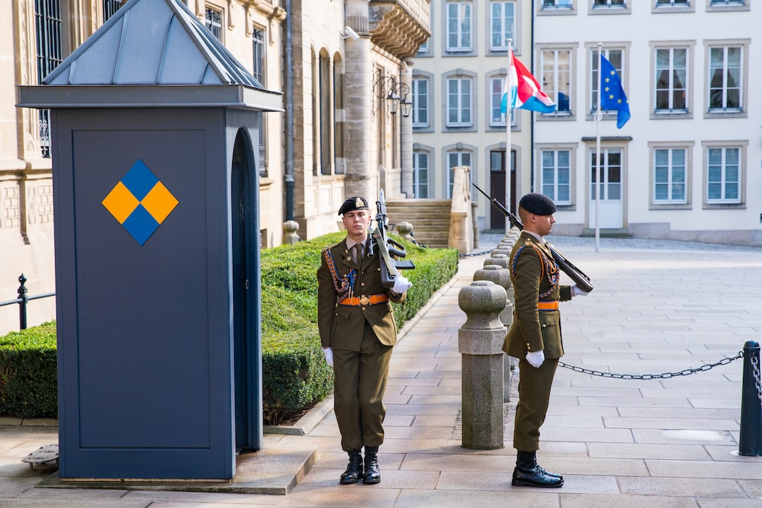 Two uniformed guards from luxury security services stand at attention outside a guardhouse near official buildings, with Luxembourg and European Union flags in the background.