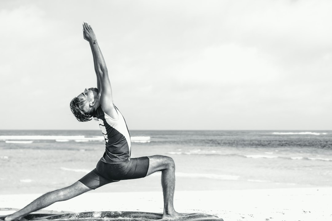 A person practices a yoga warrior pose on a sandy beach by the ocean under a cloudy sky, embodying the peaceful spirit of luxury yoga retreats in exotic locations.