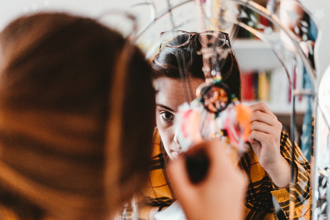 A person in a yellow plaid shirt, adjusting their hair and holding a colorful object near their face, checks their reflection—perfecting makeup for glasses and seeking make-up tips for glasses-wearing women.